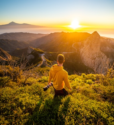 Bakifrån av mannen som håller kameran och tittar på soluppgången i bergen vid havet