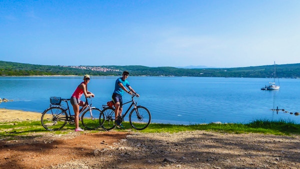 Ett par i sportiga kläder står på sina cyklar vid stranden och beundrar utsikten bakom dem. Kusten är delvis sandig, stenig och gräsbevuxen. Vattnet i viken är lugnt, båt förankrad till stranden