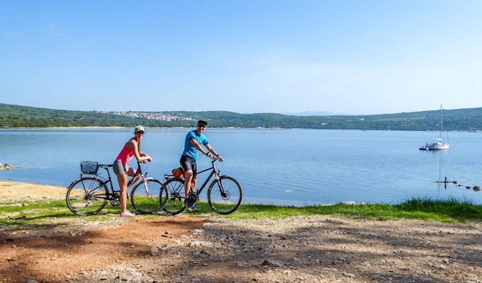 Ett par i sportiga kläder står på sina cyklar vid stranden och beundrar utsikten bakom dem. Kusten är delvis sandig, stenig och gräsbevuxen. Vattnet i viken är lugnt, båt förankrad till stranden