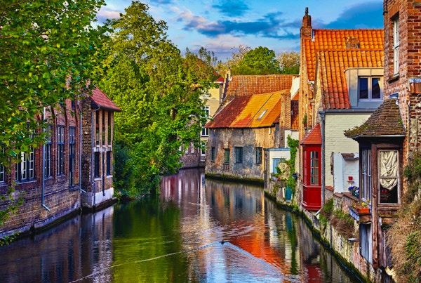 Brugge, Belgien. Medeltida forntida hus gjorda av gamla tegelstenar på vattenkanalen med båtar i gamla stan. Sommarsolnedgång med solsken och gröna träd. Pittoreska landskap.