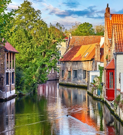 Brugge, Belgien. Medeltida forntida hus gjorda av gamla tegelstenar på vattenkanalen med båtar i gamla stan. Sommarsolnedgång med solsken och gröna träd. Pittoreska landskap.