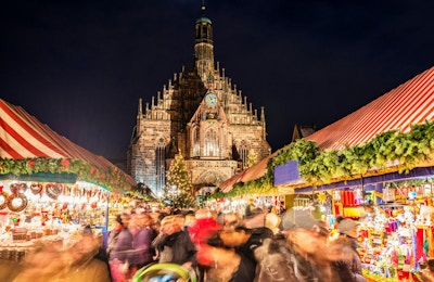 Huge crowd of people moving over NurembergÂ´s world-famous christmas market (Christkindlsmarkt) at night, passing colorful illuminated christmas decoration and food stalls. NurembergÂ´s landmark Frauenkirche (Church of our Lady) can be seen in the back.