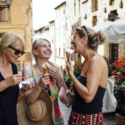 Tre mogna kvinnliga vänner som står och äter italienska glassar på en gata i Toscana under sommaren. De ler och står inför varandra och njuter av sin semester.