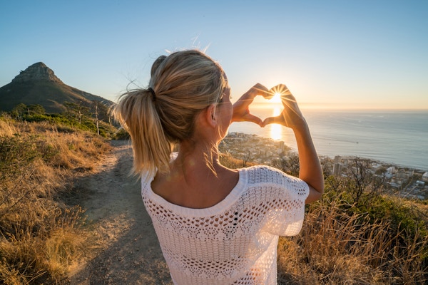 Ung kvinna vid havet som tittar på solnedgången och gör fingraramen för hjärtaform. Skott i Kapstaden, Sydafrika.
