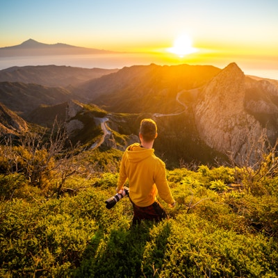 Bakifrån av mannen som håller kameran och tittar på soluppgången i bergen vid havet