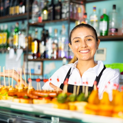 Glad latinsk kvinnlig bartender i uniform som serverar läckra tapas på spett vid disken i baren